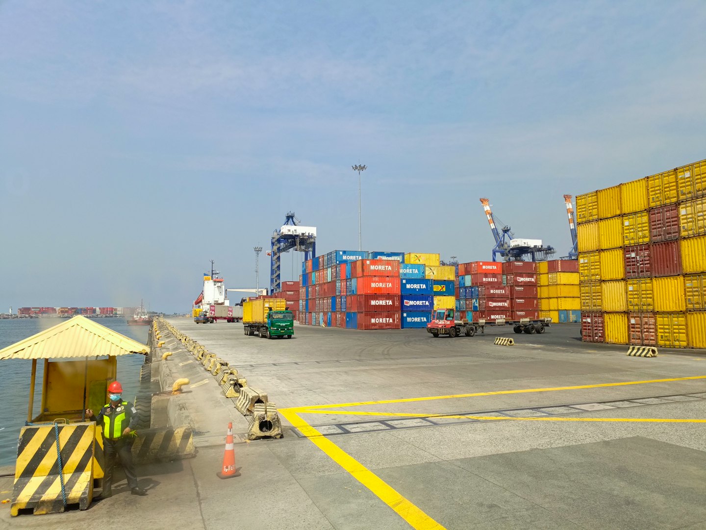 A worker in a safety vest stands at a busy shipping port.