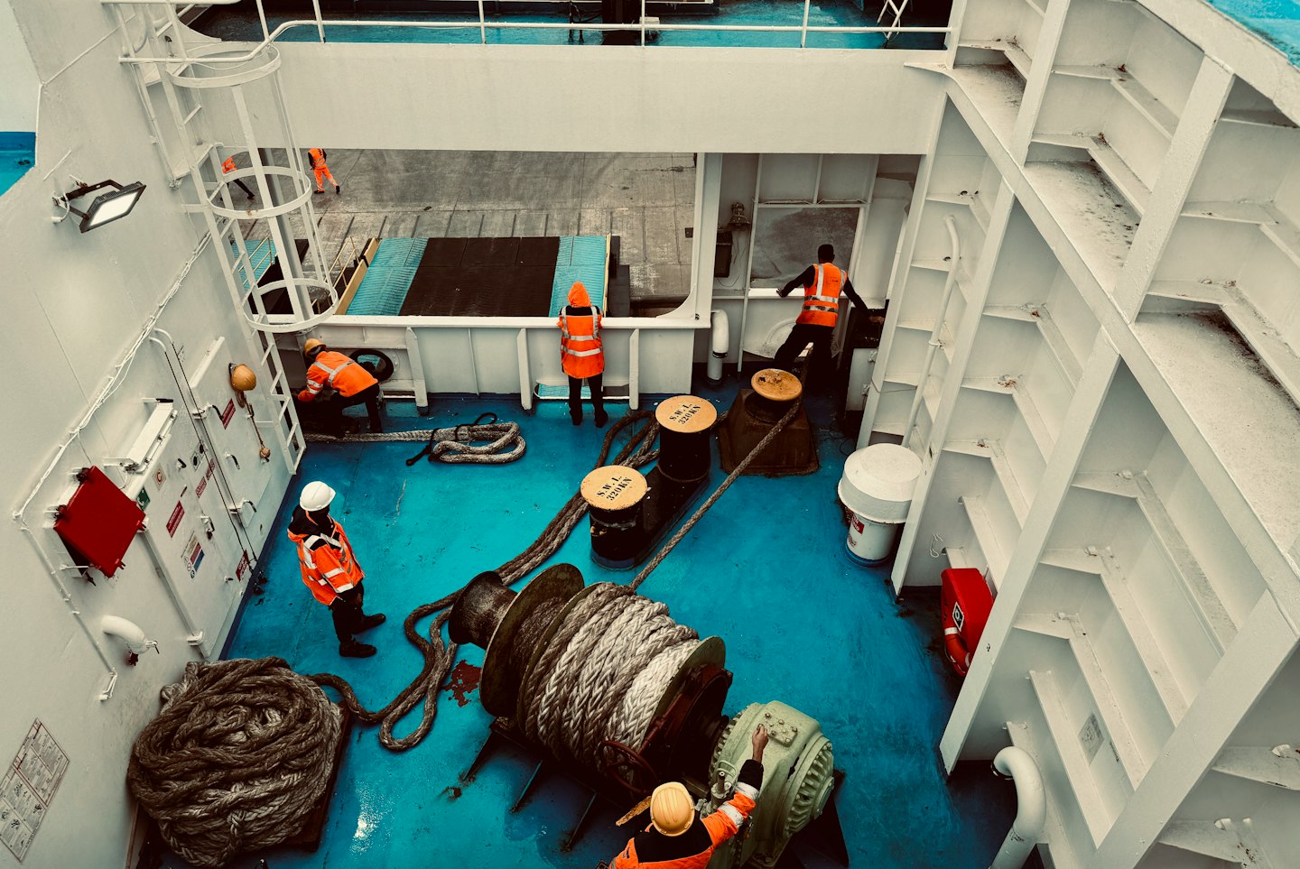 Workers in orange jackets manage ropes on a ship deck.