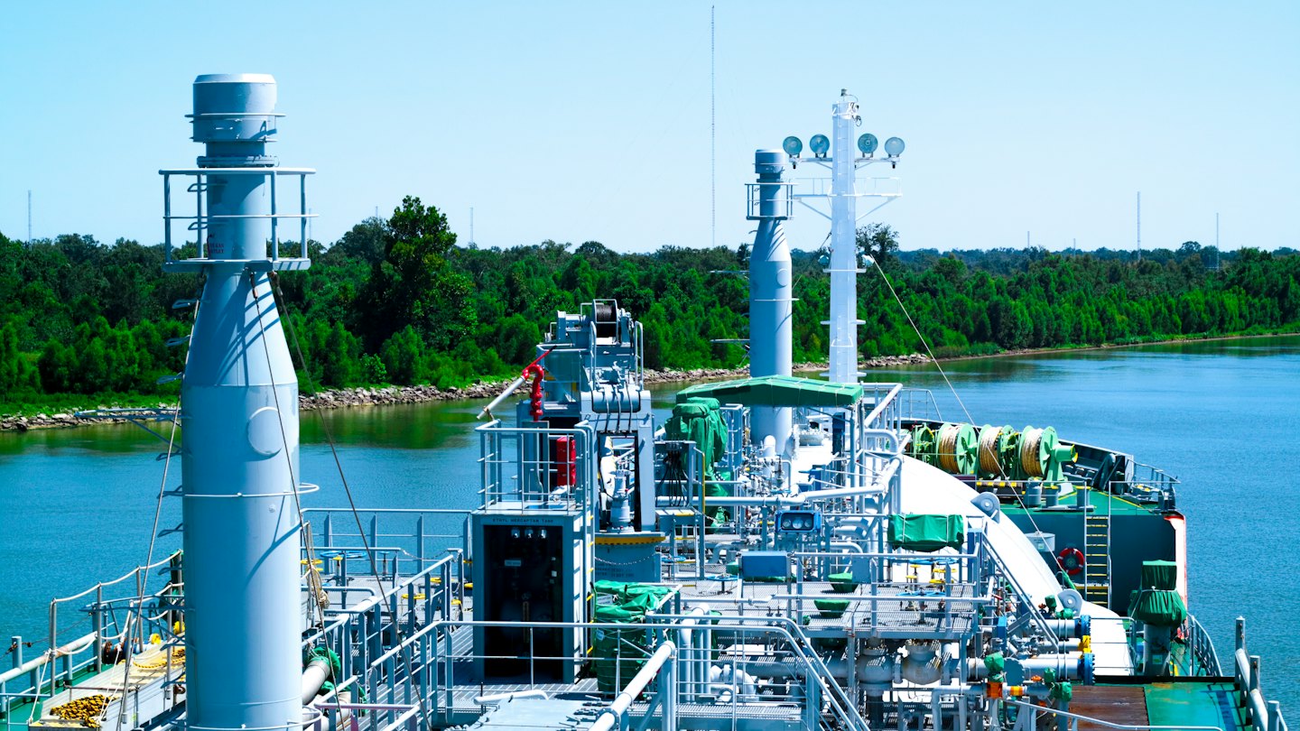 An industrial ship travels along a river past forested banks.