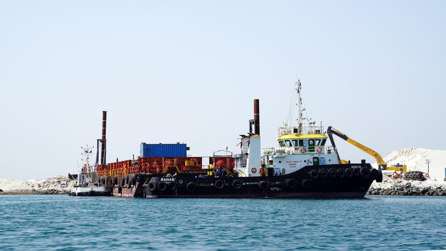 A large industrial work vessel with a tugboat on calm water.